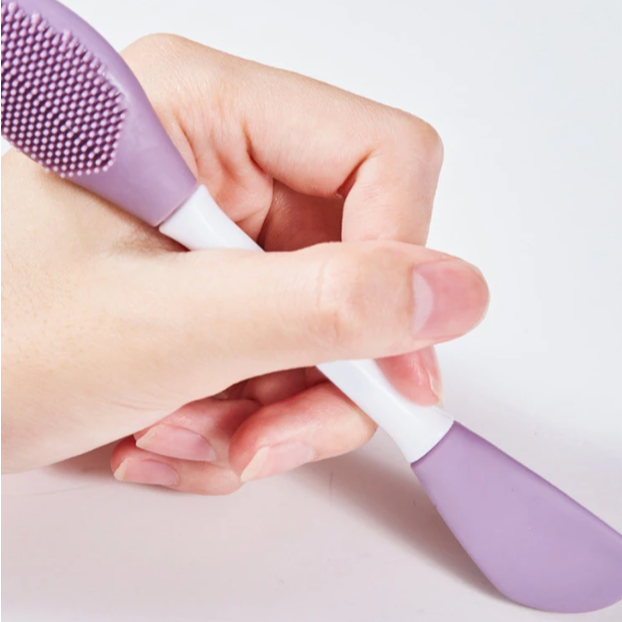 Hand holding a purple and white silicone face brush on a light background